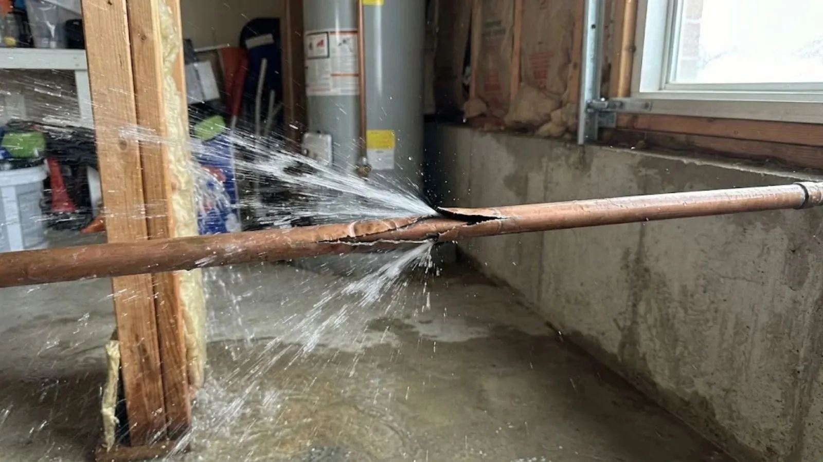 Copper pipe spraying water from a burst section beside a flooded basement wall